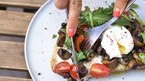 Woman hands cut canape with pouched egg mushrooms tomatoes and ruccola on piece of bread with knife and fork extreme closeup