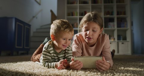 Cinematic shot of toddler baby boy and his bigger sister watching cartoons or playing games together with tablet on carpet in living room at home. technology, new generation, family.learning