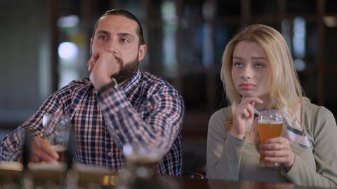 Bored girlfriend yawning sitting in pub with boyfriend watching football soccer match talking. Portrait of disinterested Caucasian young woman dating with enthusiastic man sports fan in bar indoors