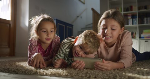 Cinematic shot of toddler baby boy and his bigger sisters watching cartoons or playing games together with smart phone on carpet in living room at home. Concept of technology, new generation, family.