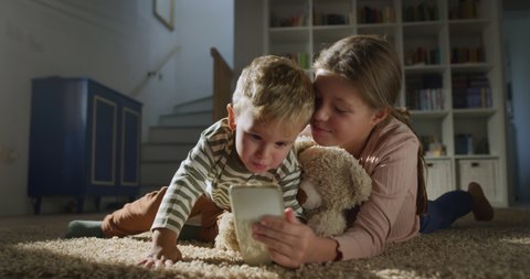 Cinematic shot of toddler baby boy and his bigger sister watching cartoons or playing games together with smart phone on carpet in living room at home. Concept of technology, new generation, family.