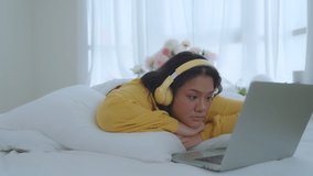 A fat woman in a yellow shirt and headphones lies in bed watching a movie in her bedroom. She shows shock or surprise while watching the movie. Holiday activities lifestyle concept.