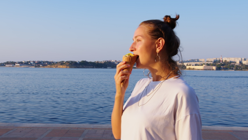 Girl walks along sea embankment, eats ice cream in wafer cone. Seaside promenade, maritime town. Young woman in silver hoop earrings, chains going along embankment, enjoying gelato in waffle cup