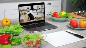 Laptop computer with man chef in screen monitor tells removes vegetables from paper bag stands on kitchen table near food ingredients and cutting board with knife, online video call cooking course