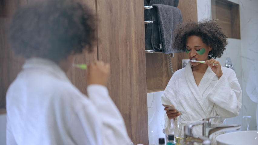 Busy woman in eye patches brushing teeth and scrolling smartphone in bathroom
