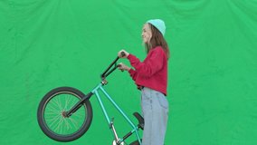 Sporty girl wearing sweatshirt and knitted hat holding bmx bike on rear wheel over green background. Pretty young woman with bicycle in her hands rotate. Chroma key. 4k raw video footage