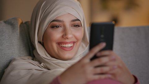 Digital tv in pocket. Close up portrait of young cheerful muslim woman in hijab watching funny movie on smartphone and laughing, spending evening at home, slow motion