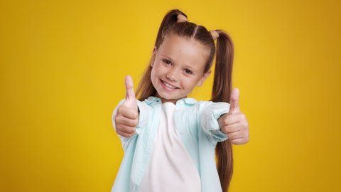 Like and approval. Studio portrait of cute little girl with two ponytails gesturing thumbs up with both hands and smiling to camera, orange background, slow motion