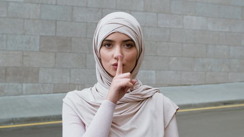 Privacy and female secrets. Outdoor portrait of young confident muslim woman wearing hijab putting finger on lips, showing shh gesture, slow motion