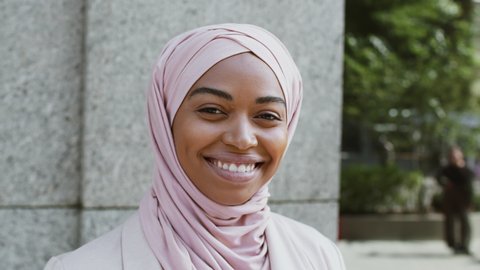 Close up portrait of happy positive muslim african american business woman wearing hijab smiling widely to camera, posing outdoors, tracking shot, slow motion