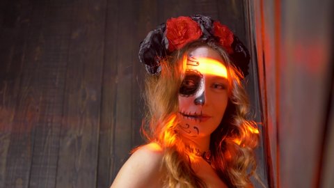 Portrait of a daring woman with Halloween makeup on her face and a hairstyle of black and red roses in red rays of light and smoke shows the middle finger close-up