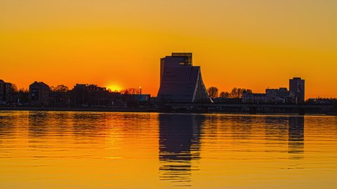 National Library of Latvia building in Riga city, reflected on Daugava river surface. Timelapse of orange sunset, with cityscape silhouettes
