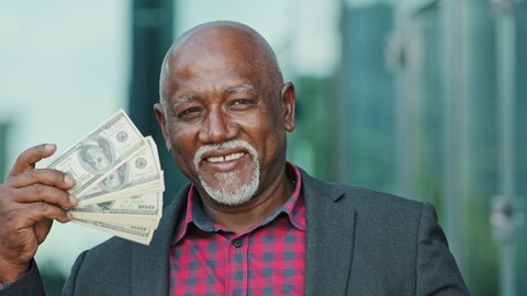 Elderly happy senior pensioner, retiree, grandfather, looking at camera, holds dollars in hands, waves fan of money to face, confident old African American man feels success, won lottery got income