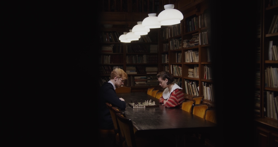 Young man and woman of European appearance start a chess game, thinking intently, sitting opposite each other at a table in the library