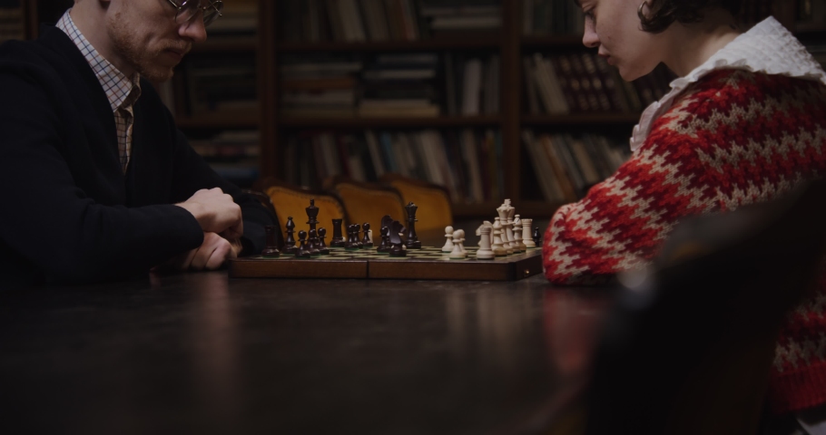 Young man and woman of European appearance playing a chess game, thinking intently, sitting opposite each other at a table in the library