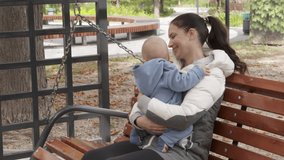 Happy family outdoors, mother with her baby sits on wooden swinging park bench enjoying autumn day. High quality 4k footage