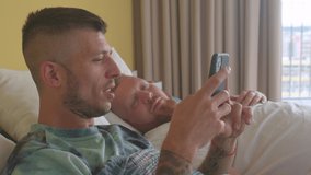 Morning in bedroom happy family reading LGBTQ+ news. Happy young gay man with his lover uses phone, sharing funny memes. Sharing moments of love and happiness. A personalized digital assistant used by