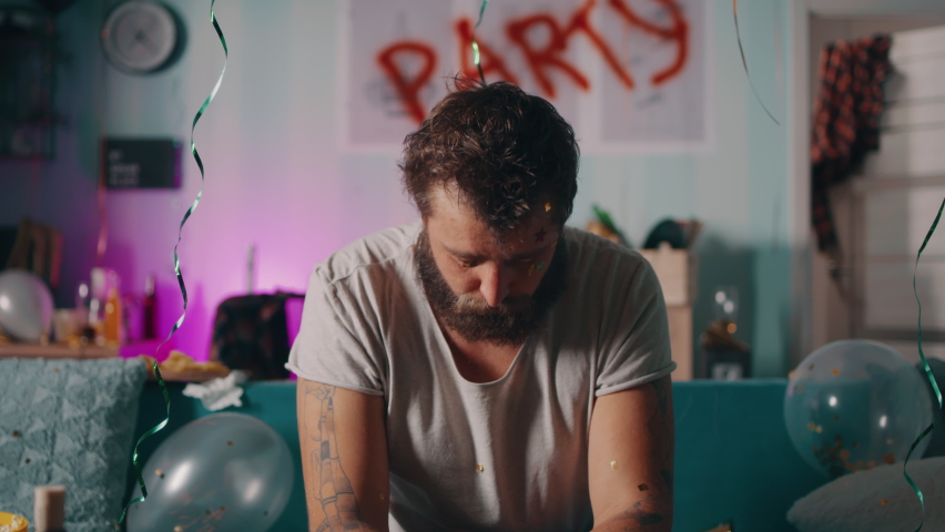 Bearded guy with hangover looking at camera and trying to spit out confetti while sitting on couch in trashed living room after party Royalty-Free Stock Footage #1080539912