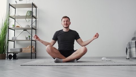 Catch zen. Relaxed man. Meditation practice. Home sport. Smiling inspired guy doing deep breath sitting yoga pose namaste hands on carpet floor in light room interior.