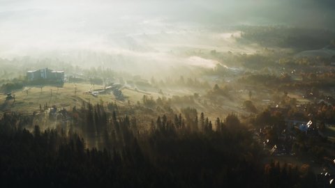 Aerial flight over Zakopane village covered with clouds of fog lit with warm morning sunlight. Drone shot mountain settlement haze sunrise light. Picturesque highland landscape. Conifers on hills
