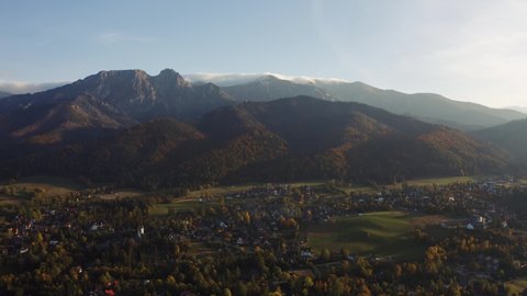 Aerial view flight over mountain village lit with warm sunlight. Drone shot beautiful highland valley in sunset light. Picturesque Tatras mountains range covered with clouds, Poland, Zakopane