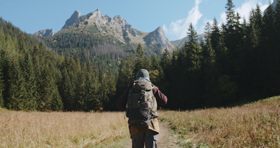 Back view handheld following male hiker walking on trail in valley with backpack, beautiful mountain ranges and forest on background. Man backpacker hiking in mountains. Adventure travel explorer Royalty-Free Stock Footage #1080542432