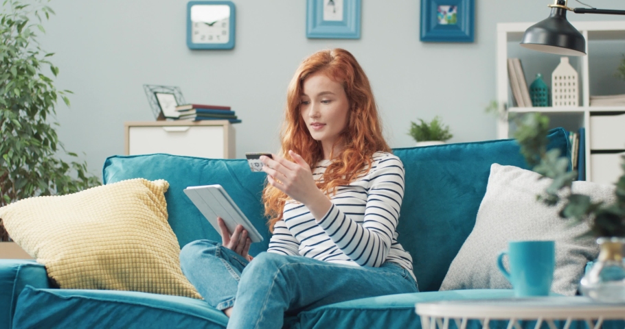 Young beautiful redhead girl with ease doing online shopping using bank card and tablet sitting at home on sofa.