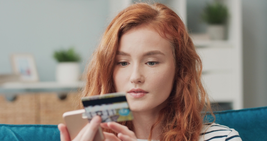 Close up of young beautiful red-haired girl enters bank card number on smartphone comparing numbers. Concept of the modern online buyer. Royalty-Free Stock Footage #1080545993