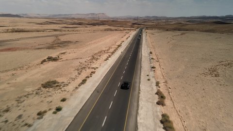 black car driving on asphalt road through the desert sands of the blue sky, white clouds, the camera flies over the land, the landscape in Israel, filming from the air, dry climate, mitzpe Ramon