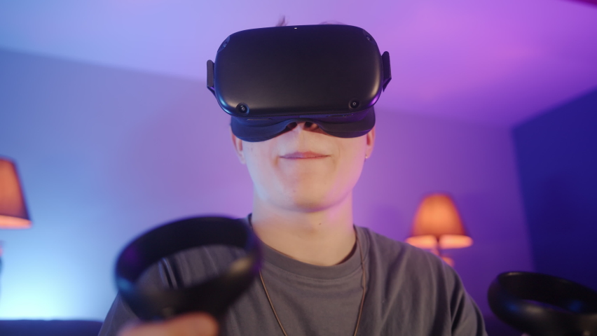 Handsome young man sitting on couch and wearing virtual reality headset and holding controllers. Active man playing video game with VR at home. He takes off the VR after finished the game Royalty-Free Stock Footage #1080550328