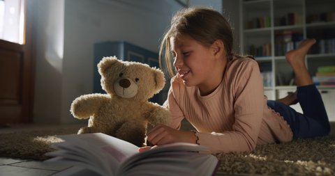 Cinematic authentic shot of cute little girl reading magic fairy tale to her teddy bear while lying comfortably on carpet in nursery. Concept of love, childhood, friendship, imagination and creativity