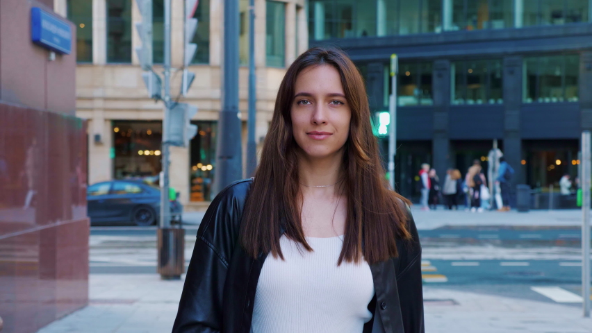 Confident businesswoman walking down street, smiling, looking at camera. Beautiful career girl in leather jacket going around city. Slender long-haired woman in hurry on business. Entrepreneur lady.