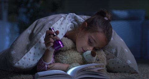 Cinematic authentic shot of little girl reading with flashlight  magic fairy tale to her teddy bear under duvet on carpet at home. Concept of love, childhood, friendship, imagination and creativity