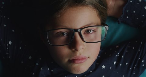 Cinematic acro shot schooler girl wearing glasses is relaxing on bed after study hard and looking in camera with soft atmospheric light. Concept of children, optics, education, study, comfort.
