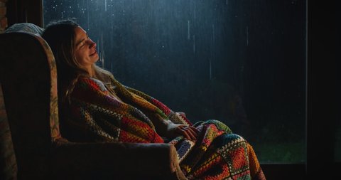 Cinematic shot of woman with colorful plaid sits in comfortable armchair and warming up hands by fireplace, relaxing while outside is raining.