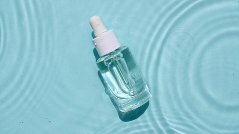 Glass cosmetic bottle with pipette lies on the surface of the water. Top view of drop falls into water and diverging circles of water on blue background. Sample of packaging for design, advertising.4k
