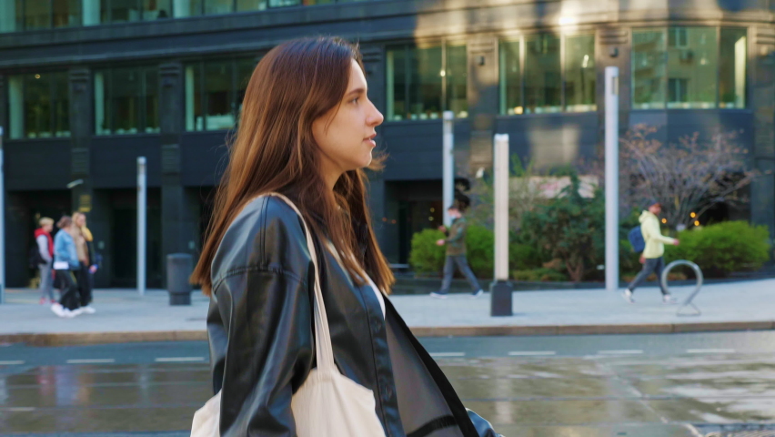 Confident businesswoman walking down street, smiling. Beautiful career girl in leather jacket going around city. Slender long-haired woman in hurry on business. Entrepreneur lady