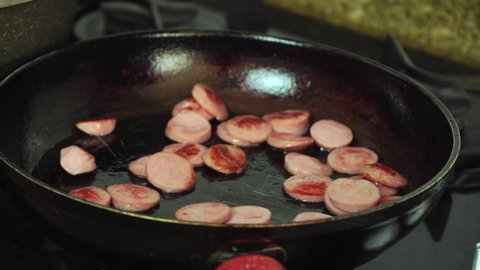 Sliced sausages are fried in boiling oil in a frying pan. A woman's hand adds red chopped bell pepper from a wooden chopping board. Preparation of snacks.