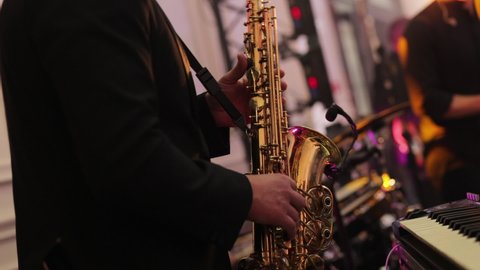 Musical band group playing song, performing on concert musician stage with lights. Saxophone player playing a solo in jazz band, performing on lightened stage. Sax musician player with band at concert
