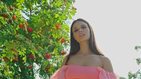 Attractive young woman looking at camera and moving elegant against trees in a park, forest or garden. Girl with naked shoulders stands under a rowan tree with clusters of orange fruits.