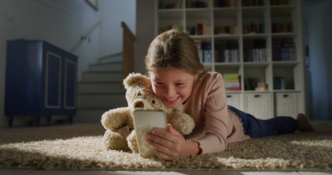 Cinematic authentic shot of cute little girl using smartphone with her teddy bear while lying comfortably on carpet in nursery. Concept of technology, childhood, friendship, imagination and creativity