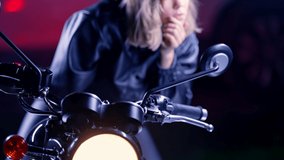 Stylish motorcyclist woman sitting on vintage-styled motorcycle and looking to mirror, corrects make-up. Young female driver at night on roadway. Trip, freedom, classic motorbike concept.