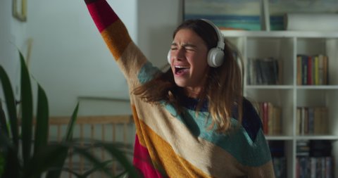 Cinematic shot of young carefree happy attractive woman with headphones is having fun to listen her favorite music playlist with headphones and dancing crazy in living room with daylight at home.