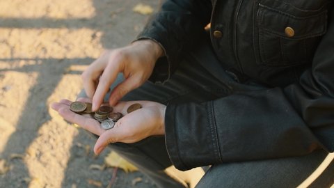 A poor man in old clothes and glasses counting coins in the palm of his hand sitting on the bench in the park. A broken person got into poverty, been laid off. Unemployment, misery concept