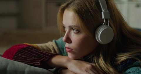 Cinematic authentic close up shot of young happy smiling woman with headphones lying on cosy carpet and relaxing with comfort while listening her favorite music playlist in living room at home.