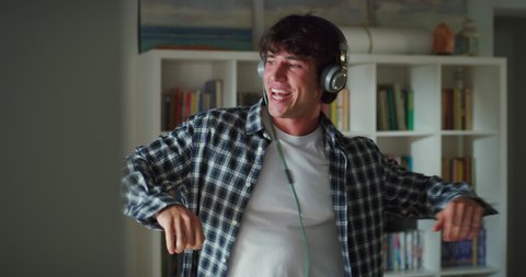 Cinematic shot of young carefree happy attractive man with headphones is having fun to listen his favorite music playlist with headphones and dancing crazy in living room with daylight at home.