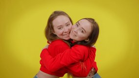 Attractive twin sisters hugging looking at camera posing with toothy smile at yellow background. Happy beautiful slim Caucasian women embracing and smiling - Powered by Shutterstock - Get 15% off with code: PIKWIZARD15
