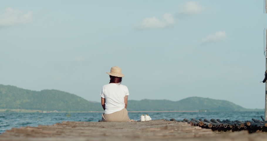 A female tourist in a brown hat, white T-shirt and brown skirt sits on a wooden bridge by the lake, looking happily at the waves, mountains and sky refreshing and relaxing on vacation.