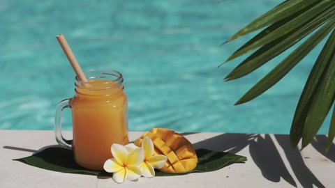 Video footage of glass mason jar with mango juice, bamboo straw, half of fresh mango, yellow frangipani flower and bubbling blue swimming pool on background.