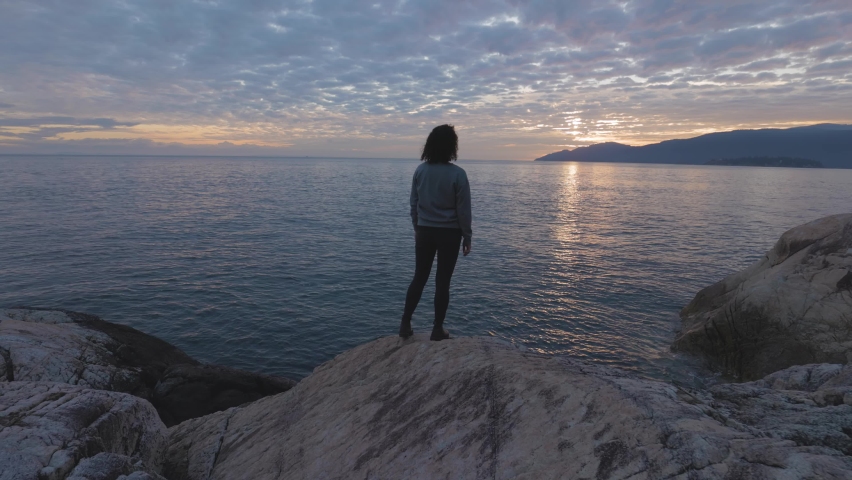 Caucasian Woman on a rocky coast during a dramatic cloudy sunset. Lighthouse Park ,Horseshoe Bay, West Vancouver, British Columbia, Canada. Adventure Travel Concept. Slow Motion
